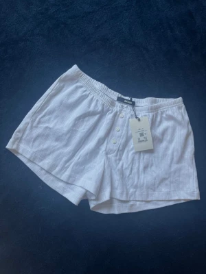 Vita shorts från BikBok - Säljer ett par vita pyjamasshorts från BikBok med diskret mönster och knappar framtill. De har resår i midjan och är tillverkade i mjukt, luftigt tyg. Perfekta för en chill och bekväm stil.