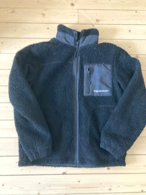 Svart teddyjacka från Jack & Jones - Mysig svart teddyjacka från Jack & Jones Originals med hög krage, hel dragkedja och bröstficka med dragkedja. Jackan har en fluffig, fårullsliknande yta och detaljer i tyg på fickan. Perfekt för kalla dagar och enkel att styla till det mesta.