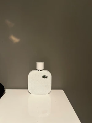 Lacoste vit parfymflaska (2/3 kvar) - Säljer en stilren parfym från Lacoste i en vit, fyrkantig flaska med rundade kanter. Flaskan har en texturerad yta och pryds av Lacostes klassiska gröna krokodillogga. Perfekt för dig som gillar minimalistisk design.