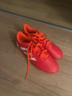 Röda Adidas fotbollsskor med dobbar - Säljer ett par röda fotbollsskor från Adidas med vita detaljer och klassiska tre ränder på sidan. Skorna har orange snörning och innersida samt dobbar för bra grepp på planen. Tillverkade i syntetmaterial och designade för att ge bra bollkontroll och stabilitet.