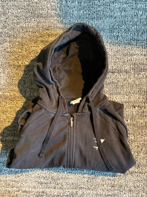 Svart zip hoodie från Gymshark - Säljer en svart hoodie från Gymshark med dragkedja framtill och två fickor. Hoodien har justerbar huva med snören och en liten vit Gymshark-logga på bröstet. Perfekt för chill eller gymmet. Materialet känns mjukt och bekvämt.  Produkten har inte ett håll men tygget saknas vid ena armen 