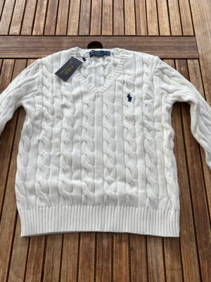 Vit kabelstickad tröja Polo Ralph Lauren - Klassisk kabelstickad tröja från Polo Ralph Lauren i krämvit färg med V-ringning och ikonisk broderad logga på bröstet. En tidlös och stilren modell som passar perfekt till både jeans och kjol.    Storlek: passar S–M.   Skick: Ny med prislapp. 