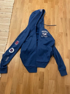 Blå hoodie från Hoodrich med tryck - Snygg blå hoodie från Hoodrich med dragkedja, huva och vita tryckdetaljer på bröstet och ena ärmen. Hoodien har känguruficka framtill och ribbade muddar. Perfekt för en streetwear-look och riktigt bekväm att bära.