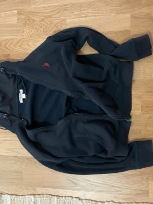 Svart hoodie från U.S. Polo Assn. - Svart hoodie med dragkedja från U.S. Polo Assn. Klassisk modell med huva och dragsko, samt röd logga broderad på bröstet. Tillverkad i mjukt material och har fickor framtill. Perfekt för en avslappnad och stilren look.