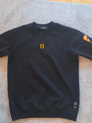 Svart Fendi sweatshirt med tryck - Cool svart sweatshirt från Fendi med gult Fendi-logo på bröstet och orange ögon-tryck på ena ärmen. Klassisk rund halsringning och ribbade muddar. Perfekt för en streetwear-look.