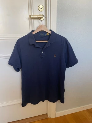 Mörkblå pikétröja från Ralph Lauren - Klassisk mörkblå pikétröja från Ralph Lauren med korta ärmar och krage. Tröjan har två knappar vid halsen och den ikoniska broderade loggan på bröstet. Perfekt till jeans eller shorts för en clean och stilren look.