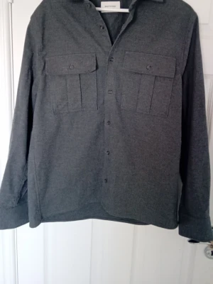 Mörkgrå overshirt från Brothers - Stilren mörkgrå overshirt från Brothers med raka linjer och två stora bröstfickor med lock. Skjortan har knäppning framtill och långa ärmar med manschett. Materialet känns mjukt och kan vara bomull eller en bomullsblandning. Perfekt lager-på-lager-plagg.