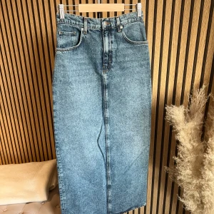 Lång jeanskjol från H&M - Trendig lång jeanskjol i klassisk blå denim med hög midja och raka linjer. Kjolen har fem fickor, bälteshällor och stängs med knapp och dragkedja framtill. Perfekt att styla med sneakers eller boots för en cool vardagslook.