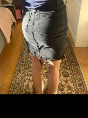 Svart jeanskjol med fransig kant - Säljer en svart, tight jeanskjol med hög midja och rå, fransig nederkant. Kjolen har klassiska fem fickor och dragkedja med knapp framtill. Perfekt för dig som gillar en edgy och avslappnad stil.