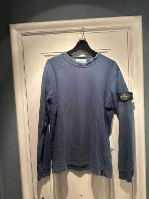 Blå långärmad tröja från Stone Island - Säljer en blå långärmad tröja från Stone Island med den klassiska loggan på vänster ärm. Tröjan har rund halsringning och är gjord i mjuk bomull.  Ett mycket litet hål på vänster ärm (se bild). I övrigt i bra skick