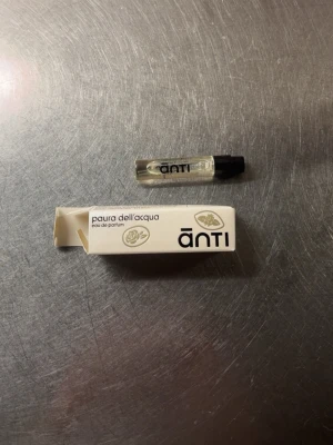 Anti Paura dell’Acqua parfymprov - Liten parfymprovflaska från Anti med doften Paura dell’Acqua. Flaskan är genomskinlig med svart lock och har en enkel, stilren design. Förpackningen är vit med svart text och dekorativa detaljer. Perfekt att testa en unik och modern doft.