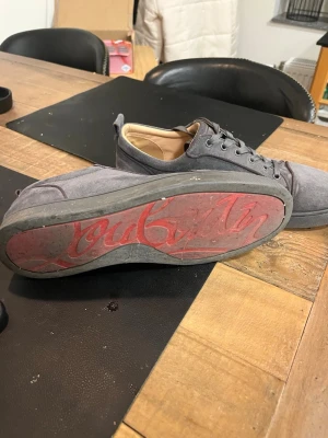 Gråa sneakers från Christian Louboutin - Tvättas innan frakt! Säljer ett par gråa sneakers från Christian Louboutin med den ikoniska röda sulan och diskret logga undertill. Skorna har lågt skaft, snörning och är tillverkade i mocka med textilfoder. Perfekta för dig som gillar stilrena och exklusiva detaljer.