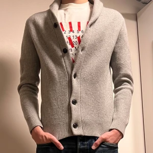 Grå ribbad kofta/cardigan från Tommy Hilfiger - storlek S/XS Säljer en stilren grå ribbad kofta från Tommy Hilfiger med knappar framtill. Koftan har en klassisk krage och långa ärmar, perfekt för kyligare dagar. Den är tillverkad i ett mjukt material som ger en bekväm passform. 95% Ull & 5% Kashmir 