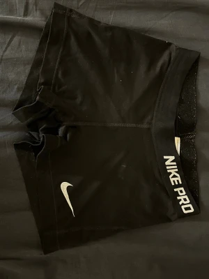 Svarta Nike Pro träningstights-shorts - Svarta Nike Pro shorts med tight passform och elastisk midja där det står 'NIKE PRO'. Perfekta för träning, tillverkade i ett stretchigt syntetmaterial som andas. Klassisk Nike Swoosh-logga framtill.