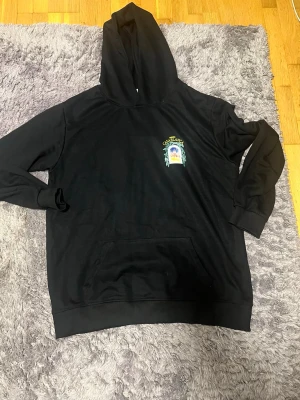 Svart hoodie Casablanca Avenida - Svart hoodie från Casablanca med tryck på bröstet och stor färgglad grafik på ryggen med texten 'Avenida Casablanca'. Klassisk känguruficka och huva. Perfekt för dig som gillar streetwear med unik design.