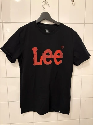 Svart Lee t-shirt med röd logga - Svart t-shirt från Lee med stor röd logotyp framtill. Klassisk passform och rund halsringning. Tillverkad i mjuk bomull, perfekt för en avslappnad stil. Snygg detalj med texturen i loggan som sticker ut.