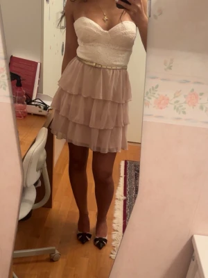 Söt axelbandslös volangklänning - Axelbandslös klänning med vit spets upptill och ljusrosa volangkjol i flera lager. Klänningen har en smal beige midjebälte.