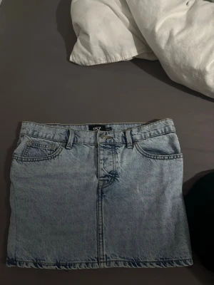 Ljusblå jeanskjol från 157 - Säljer en klassisk ljusblå jeanskjol från 157 med femficksmodell och bälteshällor. Kjolen har en rak passform och stängs med knapp och dragkedja framtill. Perfekt till sneakers eller boots för en avslappnad look.