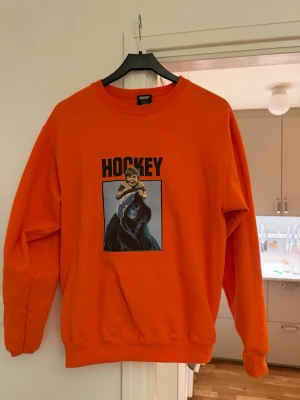 Orange sweatshirt med hockeytryck - Cool orange sweatshirt med stort tryck på framsidan som visar en björn med hockeyutrustning och texten 'HOCKEY'. Tröjan har rund hals och ribbade muddar vid ärmslut och nederkant. Perfekt för dig som gillar sportiga och unika plagg.