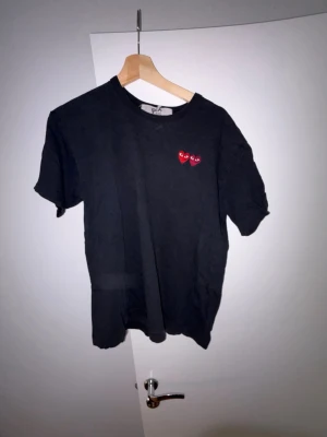 Svart Comme des Garçons PLAY t-shirt - Svart t-shirt från Comme des Garçons PLAY med två röda hjärtan broderade på bröstet. Klassisk rund hals och korta ärmar. Perfekt för dig som gillar streetwear och vill ha något stilrent men ändå med en twist. Litet hål vid märket, går lätt att sy ihop.
