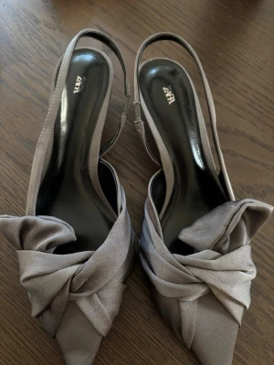 Grå satinpumps med rosett från Zara - Snygga grå pumps i satin från Zara med spetsig tå och dekorativ rosett framtill. Skorna har öppen häl med smal rem och kort klack. 