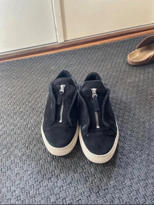 Svarta sneakers från Axel Arigato - Stilrena svarta sneakers från Axel Arigato i mocka med vit platt sula. Snygg dragkedja framtill istället för klassisk snörning ger en unik look. Låg modell med rund tå och diskret logga på sidan. Perfekta för dig som gillar minimalistisk och modern stil.