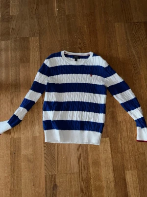 Randig tröja från Polo Ralph Lauren - Snygg kabelstickad tröja från Polo Ralph Lauren med breda blå och vita ränder. Tröjan har rund halsringning, långa ärmar och en liten röd logga broderad på bröstet. Ribbstickade muddar och nederkant ger en klassisk look. Storlek 14-16