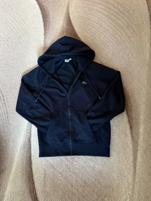 Marinblå zip-hoodie från Lacoste - Snygg marinblå hoodie med dragkedja från Lacoste. Klassisk krokodillogga på bröstet, huva med dragsko och två stora fickor framtill. Perfekt för en chill och stilren look. Mjuk bomullskänsla och ribbade muddar vid ärmslut och nederkant.