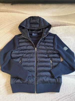 Marinblå cardigan från Aumille - Snygg marinblå cardigan från Aumille med vadderad framsida och huva. Ärmar och rygg är stickade för en cool kontrast. Jackan har två dragkedjefickor fram och en clean patch på ena ärmen. Perfekt för våren/hösten även svala sommarkvällar och riktigt trendig look.