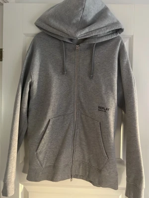 Grå zip-hoodie från Replay - Snygg grå hoodie från Replay med dragkedja framtill och två stora fickor. Tröjan är knappt använd och fräsch. Den har en stor luva och har skönt material!