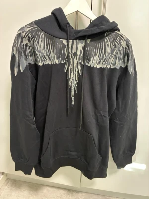 Svart hoodie Marcelo Burlon med fjädertryck - Svart hoodie från Marcelo Burlon med ikoniskt vitt och grått fjädermönster över axlar och bröst. Klassisk huva med dragsko och stor magficka framtill. Perfekt för dig som gillar streetwear med unik design.