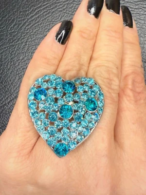 Y2K hjärtring i turkos och aqua strass – oversized statement i glam 2000-talsstil Y2K - Stor hjärtformad ring i tydlig Y2K-stil från tidigt 2000-tal, täckt av gnistrande turkosa och aqua strassstenar. Ett riktigt statement-smycke som ger maximal glamkänsla och lyfter även en enkel outfit. De olika blå tonerna skapar fint djup och lyster. Justerbar ring som passar flera storlekar.  Epok: 2000-tal (Y2K) Mått: ca 4 x 4 cm Material: silverfärgad metall, glasstrass Skick: mycket fint begagnat skick, alla stenar sitter på plats, lättare bruksspår.