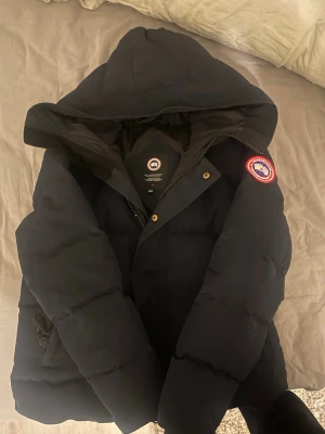 Svart dunjacka från Canada Goose - Säljer en svart dunjacka från Canada Goose med klassisk patch på ärmen. Jackan har stor huva, dragkedja och tryckknappar framtill samt flera praktiska fickor. Perfekt för kalla vinterdagar och riktigt varm tack vare dunfyllningen.