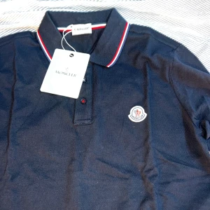 Mörkblå långärmad Moncler piké  - Snygg mörkblå långärmad t-shirt från Moncler med klassisk pikékrage och röd-vit-blå rand på kragen. Tröjan har Moncler-logga på bröstet och knappar framtill. Materialet är mjuk bomull som känns skönt mot huden. Perfekt för dig som gillar stilrena märkesplagg. 0bs lnte 0rglnal!