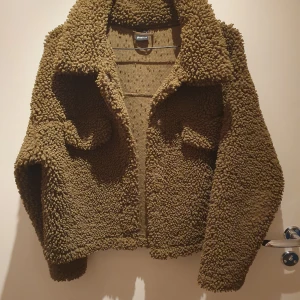 Olivgrön teddyjacka från Ginatricot - Säljer en olivgrön teddyjacka från Ginatricot med oversized passform och bred krage. Lite nopprig på insidan annars hel och ren och i mycket bra skick! 