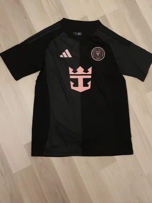 Svart Inter Miami matchtröja Adidas - Snygg svart matchtröja från Inter Miami med rosa detaljer och klubbmärke på bröstet. Tröjan har korta ärmar, rund hals och är tillverkad i ett lätt och ventilerande material. Perfekt för fotbollsträning eller att bära som supporter.