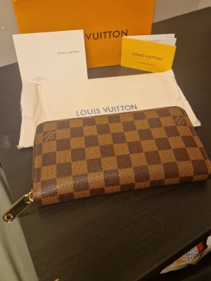 Louis Vuitton brun damier plånbok - Säljer en klassisk Louis Vuitton plånbok i brunt damier-mönster med guldfärgad dragkedja. Plånboken är tillverkad i canvas med LV-logga på framsidan och har en rektangulär form. Perfekt för dig som gillar lyxiga accessoarer.