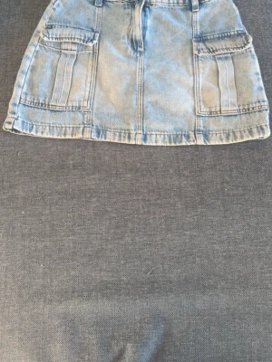 Ljusblå jeanskjol till fickor - Trendig ljusblå jeanskjol med stora fickor framtill och markerade sömmar. Kjolen har en kort passform och är perfekt för dig som gillar en avslappnad och cool stil. Snygg att matcha med sneakers eller boots.