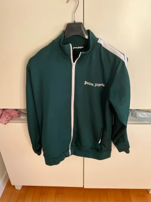 Palm Angels tröja - Mörkgrön hoodie från Palm Angels med vit dragkedja och vit rand längs ena axeln. Klassisk streetwear-look med hög krage och logga broderad på bröstet. Tillverkad i mjukt material, perfekt för en avslappnad stil.