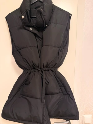 Svart lång dunväst med hög krage  - Snygg svart dunväst med hög krage och dragkedja framtill. Västen har en markerad midja med dragsko och stora fickor på sidorna. Perfekt för lager-på-lager och ger en cool siluett. Passar till många olika outfits.