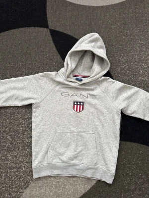 Gant hoodie - Snygg grå hoodie från GANT med broderad logga och sköld på bröstet. Klassisk känguruficka framtill och mjukt material som känns skönt mot huden. Perfekt för en avslappnad och stilren look. Strlk 158-164
