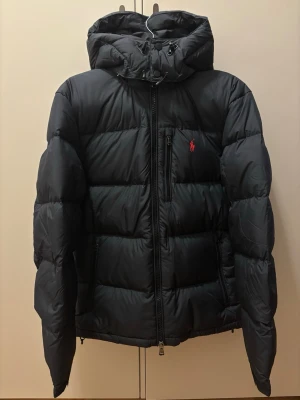 Polo Ralph Lauren - Gorham Down jacket (S) - Snygg svart dunjacka från Polo Ralph Lauren med huva och dragkedja framtill. Jackan har puffig pufferdesign, två sidofickor med dragkedja och den klassiska röda loggan broderad på bröstet. Perfekt för kalla dagar och riktigt stilren.