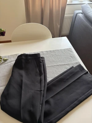 Svarta kostymbyxor Vera Petite 38 - Snygga svarta kostymbyxor från bikbok Vera Petite i modellen Low Flare. Byxorna har en klassisk passform med utsvängda ben och hällor för skärp. Perfekta för en stilren look. Materialet känns mjukt och följsamt. Aldrig använda