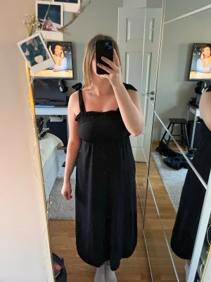 Svart långklänning med knytband - Svart långklänning med breda axelband som knyts på axlarna. Klänningen har en rak och luftig passform med rynkad detalj vid bysten. Perfekt för sommarens alla tillfällen och enkel att styla. Den har fickor på båda sidorna.