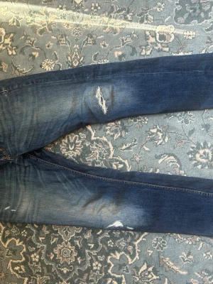 Blå slitna jeans med täckta hål - Säljer ett par blå jeans med slitningar och hål på låret för en cool, avslappnad look. Jeansen har klassisk femficksdesign och raka ben. Perfekta för dig som gillar en trendig och casual stil.