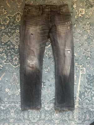 Grå slitna jeans med raka ben  - Säljer ett par grå jeans med slitningar och hål på låren och knäna. Jeansen har raka ben, fem fickor och en tvättad, lite blekt look. Perfekta för dig som gillar en avslappnad och trendig stil.