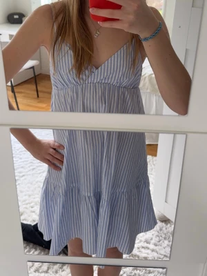Blåvit randig sommarklänning - Söt blåvit kortklänning med smala axelband och v-ringning. Klänningen är från Nelly i storlek xs men passar även s eftersom det är justerbara band och resår baktill.(Se bild) Helt nytt skick, använd en gång