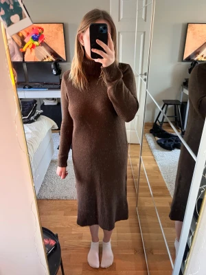 Brun stickad långklänning med polokrage - Mysig brun stickad långklänning med polokrage och långa ärmar. Klänningen har en rak passform och är perfekt för kyligare dagar. Enkel och stilren design som passar till många olika tillfällen. Liten slit på ena sidan.