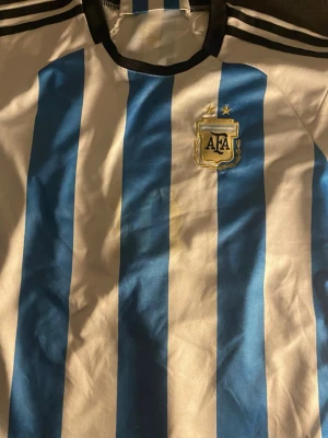 Argentina fotbollströja Adidas - Klassisk Argentina fotbollströja från Adidas med blå och vita vertikala ränder, svarta detaljer vid krage och axlar samt AFA-emblem med två stjärnor på bröstet. Perfekt för dig som älskar fotboll och vill visa ditt stöd för landslaget.