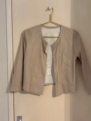 Beige kort jacka med rund hals - Stilren beige jacka med rund halsringning och trekvartsärmar. Jackan är kort i modellen och har en minimalistisk design utan synliga knappar eller dragkedja. Perfekt för att lyfta en outfit med en clean look. Materialet ser ut att vara ett tunt tyg, eventuellt bomullsblandning.